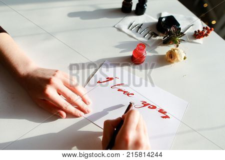 Female master of calligraphy and lettering creates red ink and pen fountain with wonderful love valentines with text I love you on day of all lovers on light table with accessories for lettering, small jars with colorful mascara and decorations in art stu