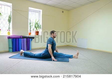 Beautiful dark-haired man performs asanas from yoga, guy sitting in room on floor on fitness mat. Inside spacious and bright, walls painted in nice yellow color, on windowsills pots with flowers. Concept of importance of keeping body in good shape, exerci