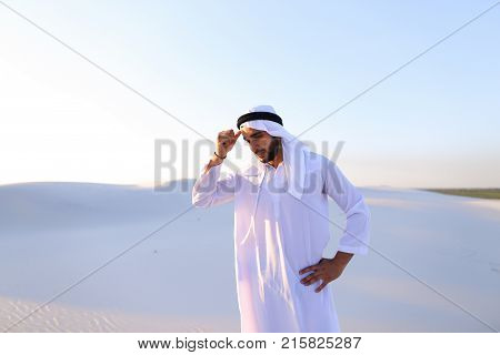 Portrait of tired Arab man, who holds head and feels migraine, tries to calm pain and massages whiskey, standing in middle of bottomless desert with snow-white sand on sunny summer day. Swarthy Muslim with short dark hair dressed in kandura, long, spaciou