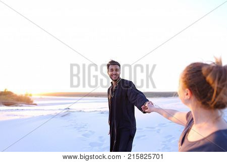 Pair of young lovers, male Muslim and woman of European appearance, look into each other's eyes and hold hands, walking along wide, bottomless sandy desert. guy and girl are smiling and laughing in open air at sunset. woman with fair hair tied in tail dre