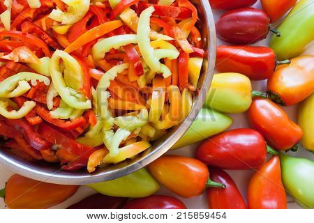 Part of large round metal bowl wiht sliced lengthwise stripes of multi-colored peppers and whole uncut peppers are lying on white table.