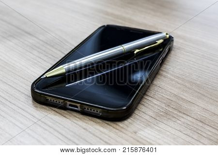 Gold-silver ballpoint pen on Black mobile phone and on wood background. Modern with a businessman.