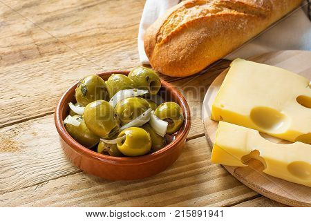 Big Spanish Green Gordal Olives with Herbs and Onions in Earthenware Bowl Baguette Maasdam Cheese on Cutting Board Wood Table Rustic Interior Mediterranean Healthy Diet Cuisine