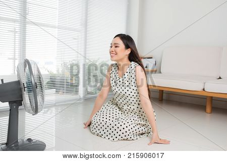 Beautiful Aisan Woman Sitting On Living Room Floor Enjoying Electric Fan Cool Wind.