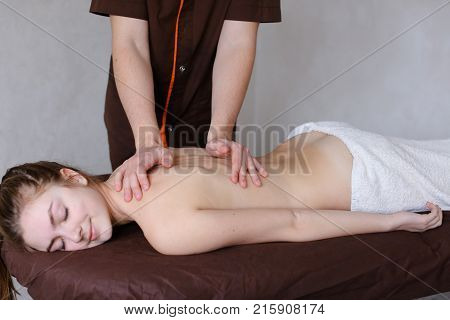 Friendly young man masseur concentrated and gently makes useful medical procedure and drives hands along back to nice girl who came to reception and lies on couch to half covered by terry towel in light cosmetology salon. Man of European appearance with s