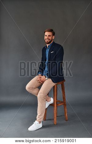 Amazing look! Full length of handsome young smiling man looking at camera while sitting on stool against grey background