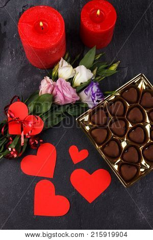 A bouquet of Japanese roses tied with a ribbon and a box of chocolates and carved hearts, burning candles on a stone background. View from above