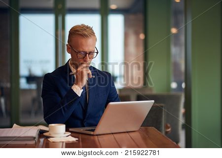 Pensive young businessman in glassses reading information on laptop screen
