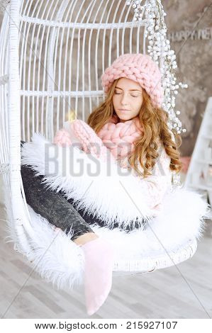 Young woman with red hair and dressed in sweater and knitted pink hat and snood sitting in her favourite chair