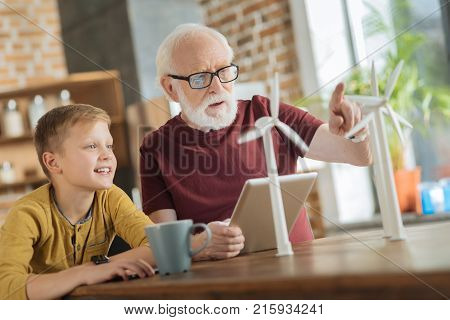 Sharing knowledge. Nice wise elderly man pointing at the windmill model and talking to his grandson while sharing his knowledge