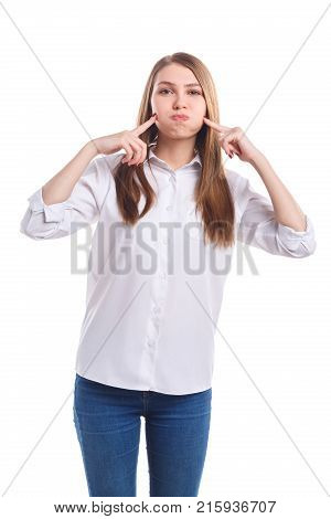 A young, cute girl in a white shirt presses her fingers on her puffed cheeks against a white isolated background