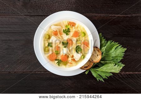 An overhead photo of a plate of chicken, vegetables, and noodles soup, shot from above on a dark rustic texture with slices of bread, a celery branch, and a place for text