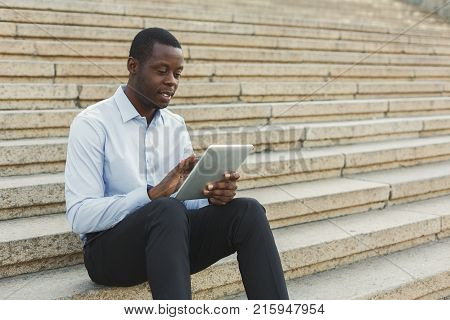 Happy black businessman working on tablet. African-american insuranse agent browsing news on digital device, sitting on the stairs outdoors in urban area