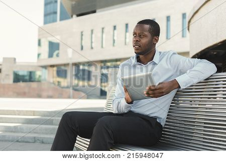 Pensive black businessman working on tablet. African-american insuranse agent browsing news on digital device, sitting on the bench outdoors near modern office center in urban area cityscape