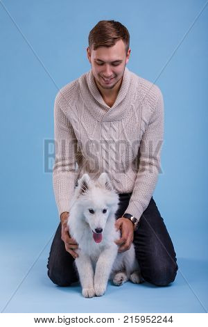 A young European guy sits and plays with a cute Samoyed puppy. On a blue background.