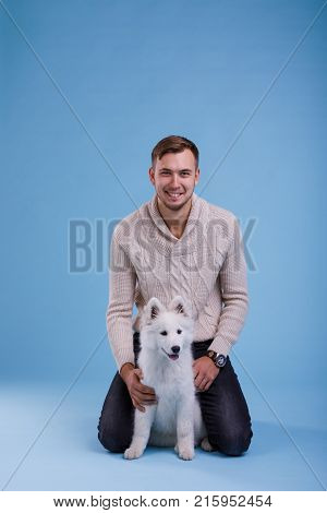 A young happy European guy sits and plays with a cute little Samoyed puppy. On a blue background.