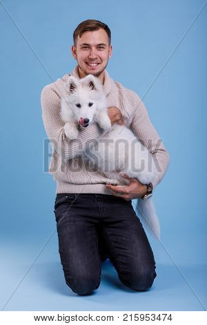 A European young brunette guy is sitting and holding a small cute little puppy Samoyed and smiling cute. On a blue background.
