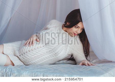 Beautiful Pregnant Woman lying in bed with canopy. Wears white knitted pullover. Maternity concept.