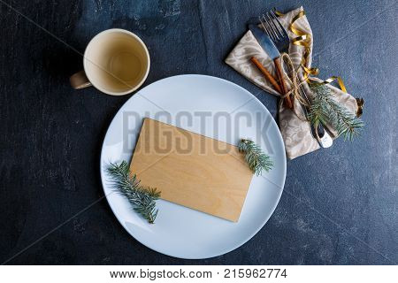 On a dark surface, a white round plate with a wooden veneer inside, next to the cutlery wrapped in a tissue serving napkin and decorated with cinnamon sticks and twigs of a Christmas tree and next to an empty cup. View from above.