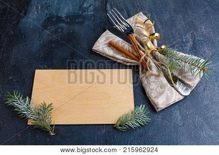 On a dark, worn surface, an empty plywood board with Christmas trees and next to a cutlery wrapped in a tissue serving napkin and decorated with fragrant cinnamon sticks and small twigs of Christmas trees.