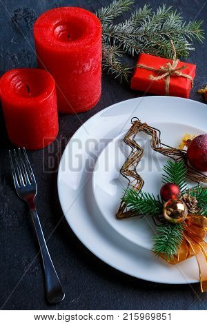 On the table is a round white plate with a decorative Christmas tree with ribbon and Christmas balls, next cutlery, red candles and a small red gift box with a sprig of Christmas tree.