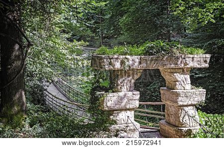 Old suspension bridge based on stone pillars across the stream in the arboretum.