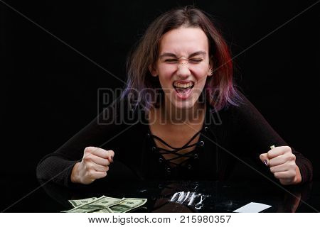 A European girl drug addict in a black sweater and with violet hair angrily screams sitting at the table with a dosed cocaine next to the Dolar bills. On a black background.