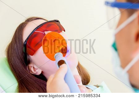 Young man male dentist in medical glasses and mask treatments teeth of woman female patient in red uv glasses by uv equipment tool implanting medical sealants