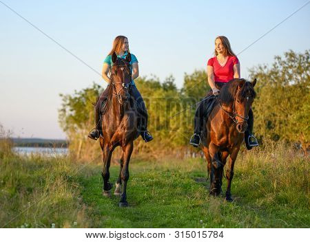 Two Happy Teenage Girls Are Astride Their Bay Horses In Rural In A Summer Evening.