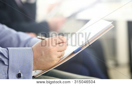 Focus On Male Hand Writing Something. Businessman Sitting In Modern Conference Room And Holding Pape