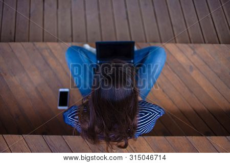 The Top Of The Head Is A Brunette Student Girl With Long Flowing Hair Sitting Cross-legged, On Woode
