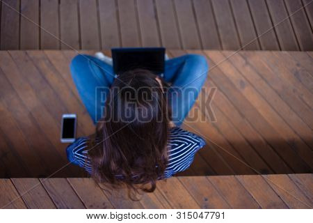 The Top Of The Head Is A Brunette Student Girl With Long Flowing Hair Sitting Cross-legged, On Woode
