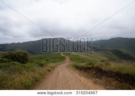 Long, Winding Hiking Trail Leading To Mountains With Two Hikers Marching Into The Distance