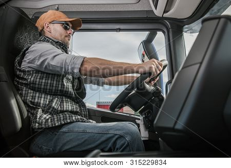 Trucker On The Route To Destination. Cargo Shipping Theme. Caucasian Semi Truck Driver Inside Vehicl