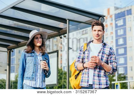 Happy Couple Hipster Travelers With Backpacks In Bus Stop With Cups Of Drinks Outdoors With Urban Ci