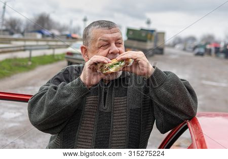 Senior Driver Gobbling Lyulya Kebab In Lavash Near His Car Standing On A Back Road