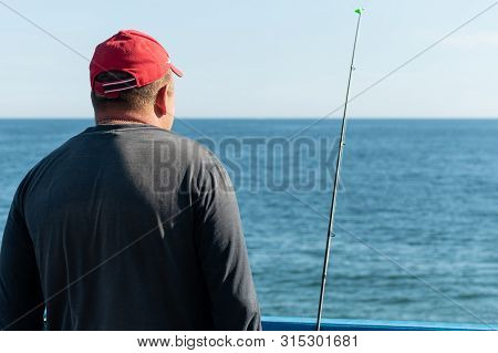 Fisherman Fishing From Pier. Man With Fishing Rod In Red Cap. Windless Weather, Calm. Copy Space. Ri