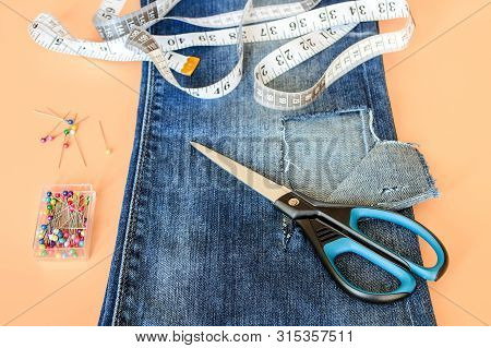 Blue Jeans With Large Hole Folded In Half, Sewing Pins In A Box, White Tailor Tape With Centimeters 