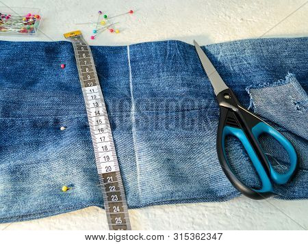 Blue Jeans With Large Hole Folded In Half, Multi Coloured Sewing Pins In A Box, White Tailor Tape Wi