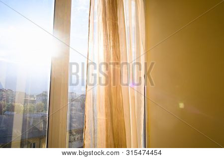 Sun Shining Through Curtains And Window. Sunlit Apartment On Sunset. City View And Blue Sky.