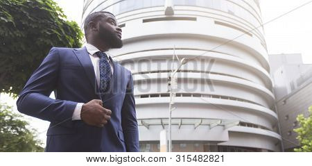Portrait Of Thougtful Confident Black Businessman Against Modern Office Center Background, Low-angle