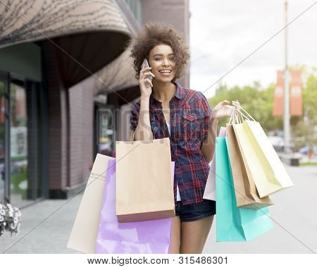 Young African American Girl Carrying Colorful Paper Bags, Talking By Cellphone, Street Background