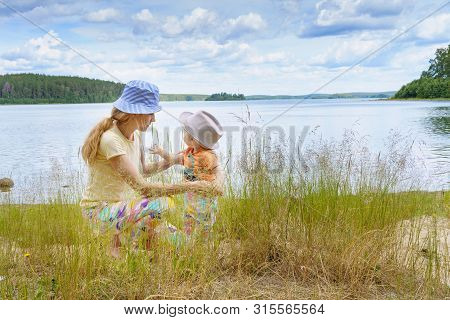 Mom And Her Little Son Walk And Communicate On The Shore Of A Forest Lake On A Summer Day Against A 
