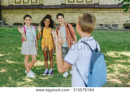 Back View Of Shoolboy Waving Hand To Smiling Multicultural Friends