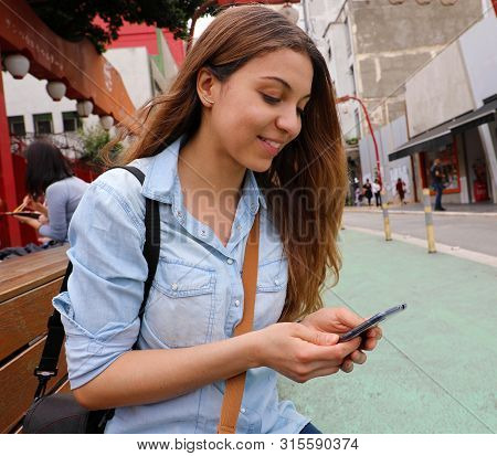 Beautiful Young Woman Sitting On Street Bench Messaging With Mobile Phone In Sao Paulo City, Brazil