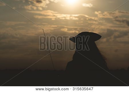 Silhouette Of Pirate Woman In A Pirate Hat And With A Sword In Hands Over A Evening Cloudy Sky Backg