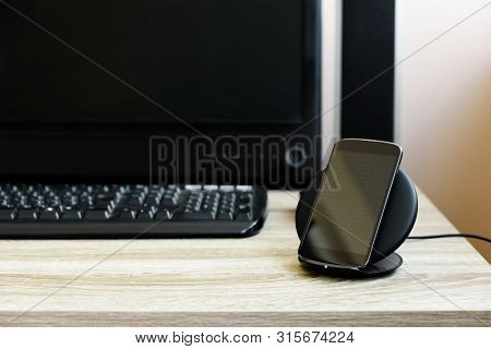 Black Smartphone Charged By Wireless Charger Against Computer Monitor, Keyboard And Speakers On Wood