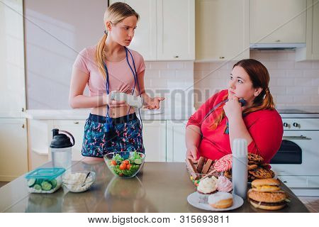 Young Slim And Overweight Women Look At Each Other In Kitchen. Thin Model Hold Bowl With Salad In Ha