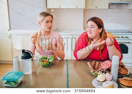 Confused Young Slim Model Eat Farmer Cheese At Table. She Look At Plus Size Model Who Eats Burgers A