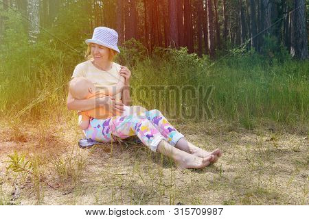 Happy Mother Holds Her Baby In Her Arms, Smiles And Breastfeeds Him On The Shore Of A Forest Lake On